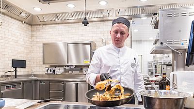 Mann in weißem Kittel und Kochmütze schwenkt eine Bratpfanne in einer Gastroküche