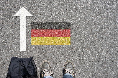 Asphalt mit einem weißen Pfeil nach oben, einer Deutschlandflagge und zwei Füßen in Schuhen