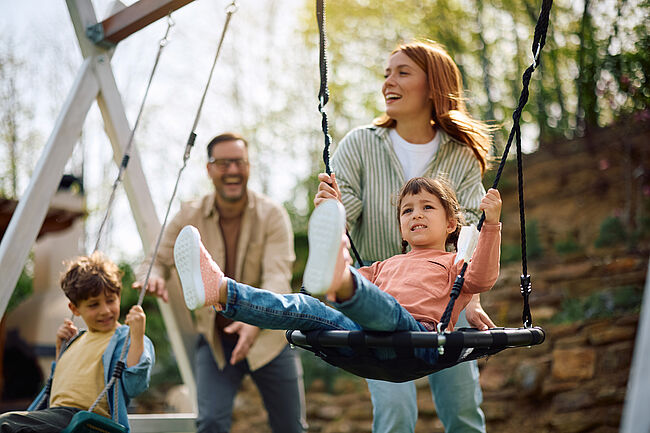 Glückliche Familie mit zwei Kindern beim gemeinsamen Spielen auf einer Schaukel im Park