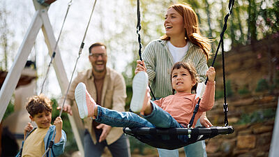 Glückliche Familie mit zwei Kindern beim gemeinsamen Spielen auf einer Schaukel im Park