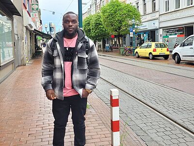 Ein junger Mann steht in einer Straße, im Hintergrund eine Straßenlaterne, Straßenbahnschienen und parkende Autos