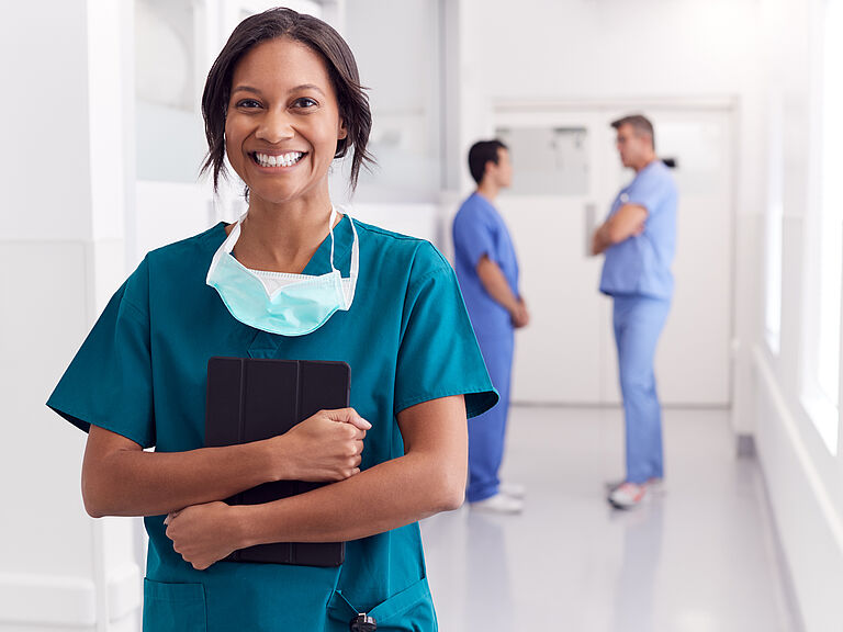 Smiling healthcare professional in scrubs holding a digital clipboard in a hospital hallway