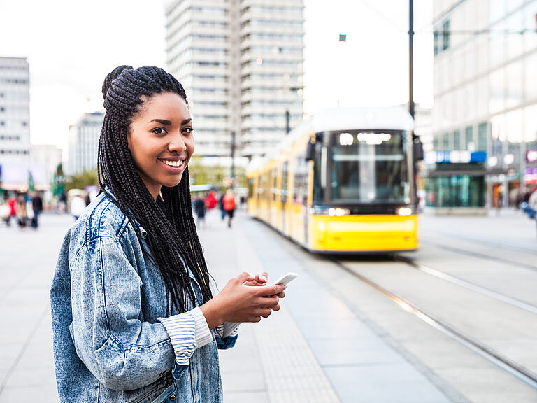 Lächelnde junge Frau mit Smartphone an einer Haltestelle vor einer gelben Straßenbahn in der Innenstadt