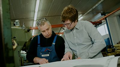 Zwei Männer in einer Werkshalle schauen konzentriert auf ein großes Blatt Papier