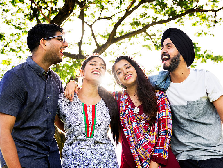 Four young adults laughing together with their arms around each other in a sunny park