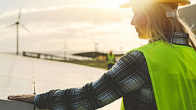 Person wearing a safety helmet and high-visibility vest looks at wind turbines at sunset – a symbol of renewable energy and sustainable jobs.