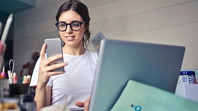 Mujer joven sentada en un escritorio con un ordenador portátil y sosteniendo un teléfono móvil en la mano.