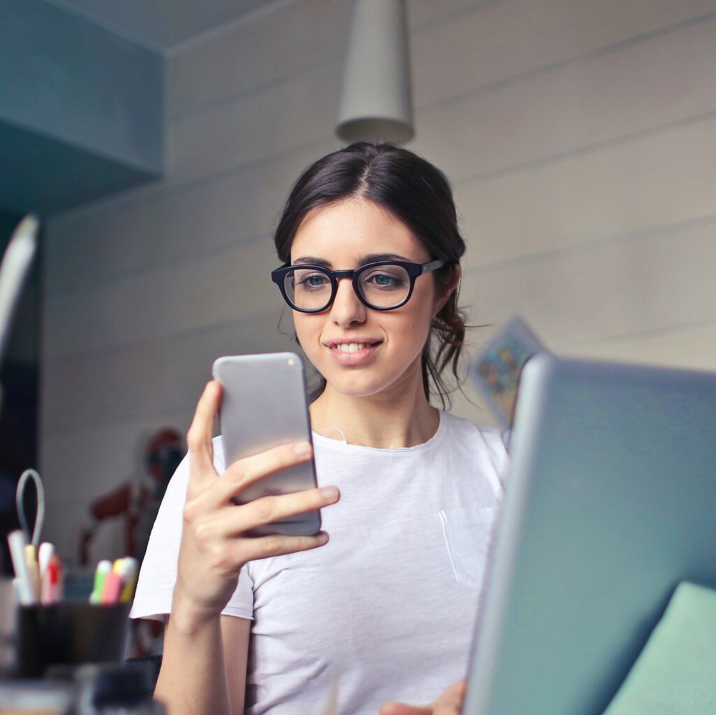 Young woman sitting at a desk with a laptop and holding a mobile phone in her hand