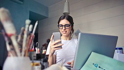 Junge Frau sitzt am Schreibtisch mit dem Laptop und hält das Handy in der Hand