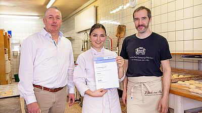 Bäckerin mit Urkunde steht zwischen zwei Bäckern