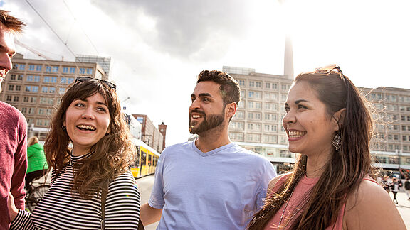 Imagen simbólica de la hermosa vida en Alemania: Tres personas están relajadas en un entorno urbano soleado con edificios modernos y un tranvía amarillo al fondo.