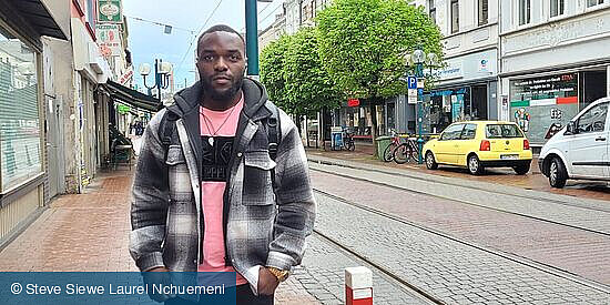 Ein junger Mann steht in einer Straße, im Hintergrund eine Straßenlaterne, Straßenbahnschienen und parkende Autos