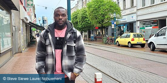 Ein junger Mann steht in einer Straße, im Hintergrund eine Straßenlaterne, Straßenbahnschienen und parkende Autos
